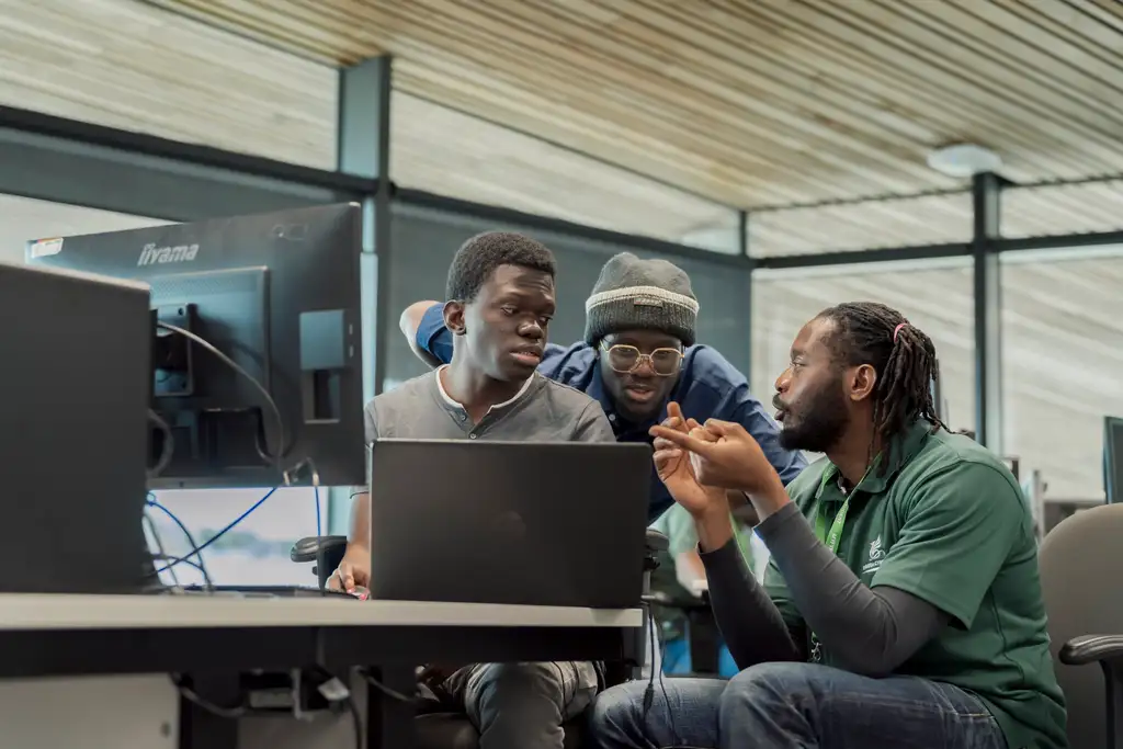 Three students of the global majority sit around a laptop and discuss their work in a cyber security workshop. They all have concentrating facial expressions