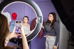 Someone is filming two fashion promotion students through a ring light as they talk about clothing.