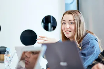 A person sat with their laptop in the Newport library talking to other people that are sat with them. You can see the reflections of other people in the screens of their devices.