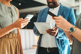 Three staff people stand together in an office on two phones and a tablet collaborating on work