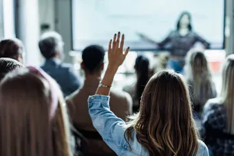 A person raises their hand while listening to a colleague's presentation in a conference room with a large screen