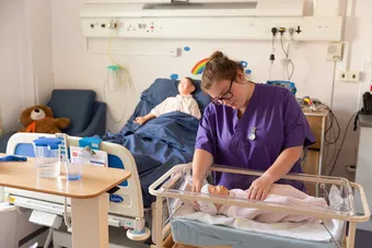 A student nurse wearing purple scrubs is caring for two mannequins that simulate a baby and a child in a simulated hospital environment.