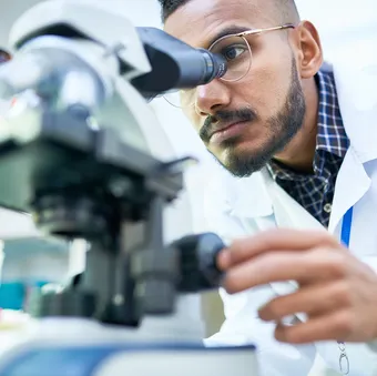 A scientist in a lab coat and glasses looks intently into a microscope