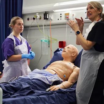 Student nurse listening to lecturer whilst treating a dummy in the clinical simulation centre