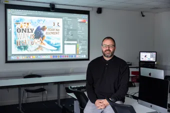a university lecturer sitting on a desk in classroom smiling to camera