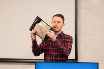 cyber security lecturer in red and black shirt holding up book in front of whiteboard