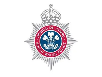 The South Wales Police logo - a silver police star style crest with the Welsh feathers in the middle in a navy blue circle and their German motto 