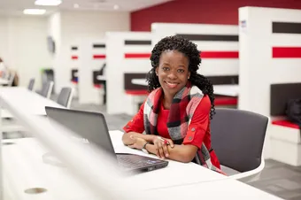 A student sat in the library working on a laptop and smiling at the camera.