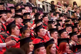 Students wearing graduation robes and caps cheer and clap for their peers during their graduation ceremony