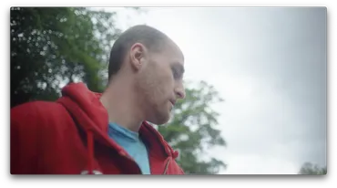 A man in a red hoodie is looking down at an angle with trees and sky visible behind him.