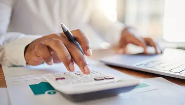 A close up of someone using a calculator and holding a pen whilst working out budgets.