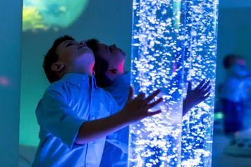 Child in therapy sensory stimulating room interacting with coloured lights bubble tube lamp during therapy session