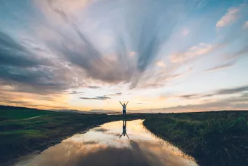 A young man holding his hands up to the sky, reflected on a puddle at sunset