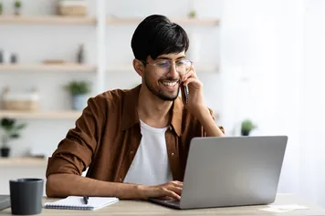 Cheerful student sitting at desk with laptop having phone conversation, typing on computer keyboard