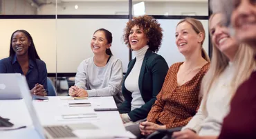 A group of women in smart casual dress sat around a table in a meeting room, smiling.