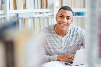 A person sitting at a desk in Newport library with their laptop, smiling at the camera. There are bookshelves in the background.