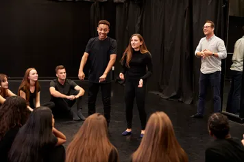 An academic watches two drama students performing for their classmates in a black drama rehearsal studio