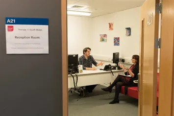 Two people sat, talking to eachother, in a therapy room. One is sat behind a desk and the other is sat on the sofa.
