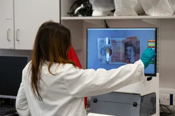 A Forensic Sciences student dressed in a white laboratory coat uses the Video Spectral Comparator to examine a £10 bank note in a USW forensic laboratory