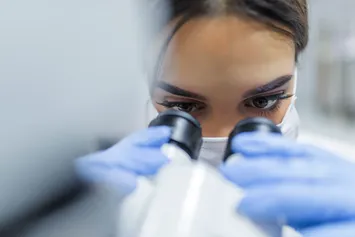 A close-up of a person wearing blue medical gloves and a face mask looking into a microscope