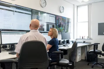 Two people observing multiple TV and computer screens showing various evidence.