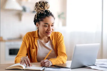student working remotely at home, sat at a laptop writing in a notebook