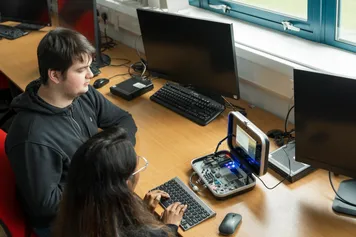 Two computing students sit at a desk using a small portable computer with flashing lights and a keyboard in a computing workshop while surrounded by larger conventional desktop monitors