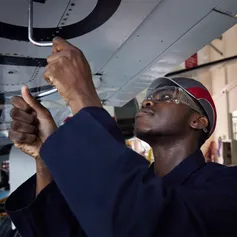 A student wearing a navy engineering boiler suit, an engineering hat and safety glasses uses a drive speeder wrench on the underneath of a wing on the small plan at the USW aerospace centre in Treforest