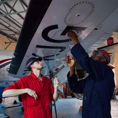 Two students in overalls carry out maintenance on the underneath of an aircraft's wing