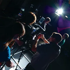 A group of students help to operate a camera filming two people chatting under a spotlight in a dark film studio