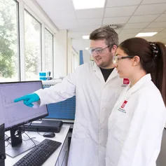 Two students in white laboratory coats and blue scientific gloves point at a computer inside the USW forensic science lab