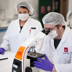 A USW forensics student looks at a specimen in a petri dish under a microscope in a USW laboratory while wearing a USW white lab coat, purple scientific gloves and a hair net