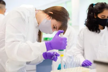 A student with long tided back hair conducts an experiment using small test tubes and a pipette while wearing a white laboratory coat, purple latex gloves and a light lilac face covering at Glyntaff Campus