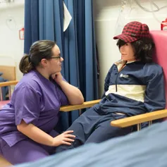 A mental health nursing student practices talking to a dummy patient in a hospital chair next to a hospital bed in a simulation ward at USW Glyntaff