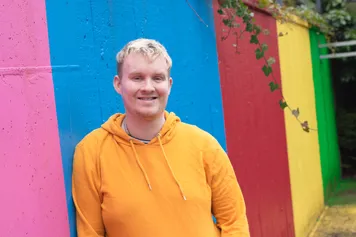 Will Simpson smiling at the camera leaning against a multi-coloured wall.