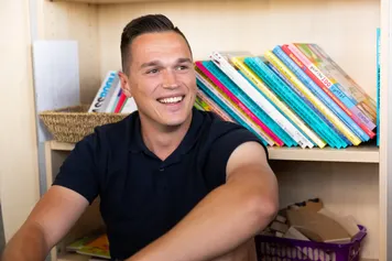 A student sat on the floor smiling towards the camera with a bookshelf behind him