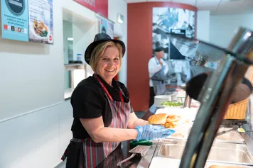 a member of University catering staff, standing behind a hot food counter serving a burger and chips