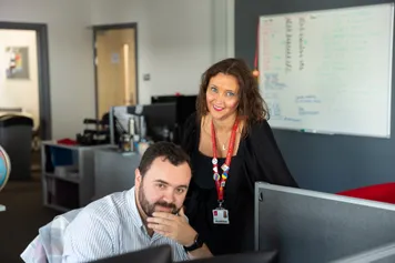 two members of University staff, one sitting at a computer and one standing, smiling to camera