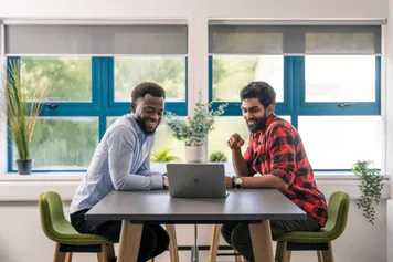 two members of university staff sat at a communal working table, looking at a laptop