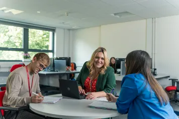 three members of the faculty of business and creative industries team sitting around a table having a meeting, taking notes in notebooks and a laptop