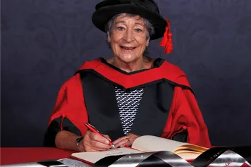 Beti George signs a book on a red desk while wearing an honorary graduate's cap and gown in front of a blue backdrop