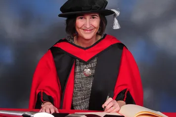 Clare Hudson smiles at the camera while signing a book on a red desk and wearing a doctorate's cap and gown in front of a blue backdrop