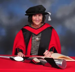 Clare Hudson smiling at the camera while wearing a PhD graduates cap and gown and writing in a book at a red desk with the celebratory mace in front of her