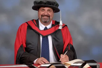 Jonathan Crego smiles at a camera while signing a book on a red desk and wearing a doctorate's cap and gown in front of a blue backdrop