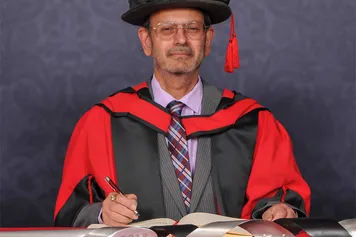 Keshav Singhal looks at the camera ands signs a book on a red desk while wearing a doctorate's cap and gown in front of a blue backdrop