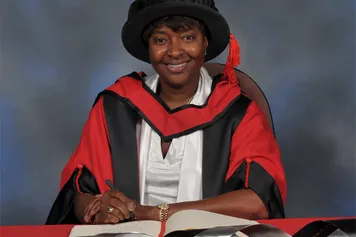 La-Chun Lindsay smiles at the camera and writes in a book on a red desk while waring a PhD cap and gown in front of a blue backdrop