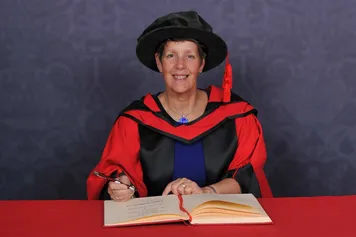 Pippa Britton writes in a large book on a red desk and smiles at the camera while wearing a doctorate's cap and robe in front of a blue backdrop