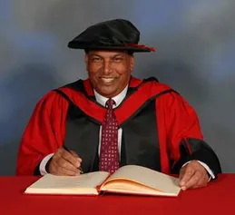 Raj Aggarwal smiling at the camera while wearing a doctorate's gown and cap and writing in a book on a red desk in front of a blue backdrop