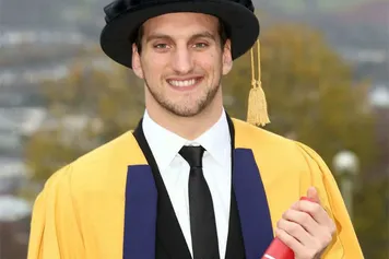 Sam Warburton smiles at the camera and holds their degree outside while wearing a doctorate cap and gown