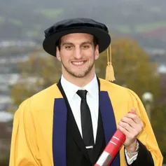 Sam Warburton smiles at the camera and holds their degree outside while wearing a doctorate cap and gown